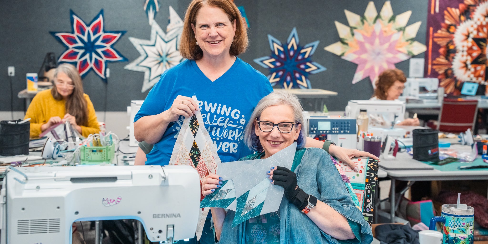 Quilters at a Stitchin’ Heaven sewing retreat hold up pieced quilt blocks, with finished star quilts on the wall and others sewing at machines behind them.
