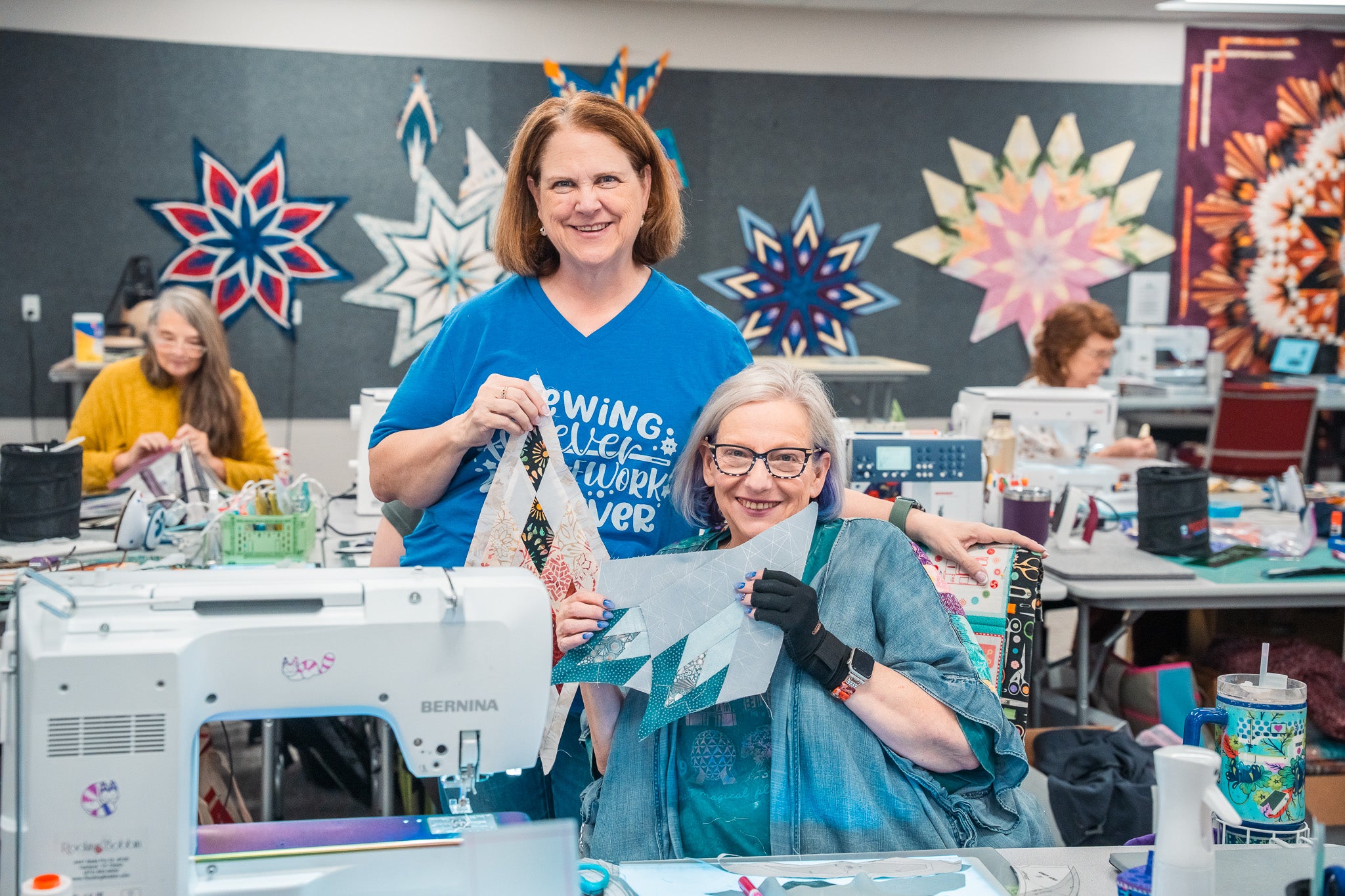 Quilters at a Stitchin’ Heaven sewing retreat hold up pieced quilt blocks, with finished star quilts on the wall and others sewing at machines behind them.